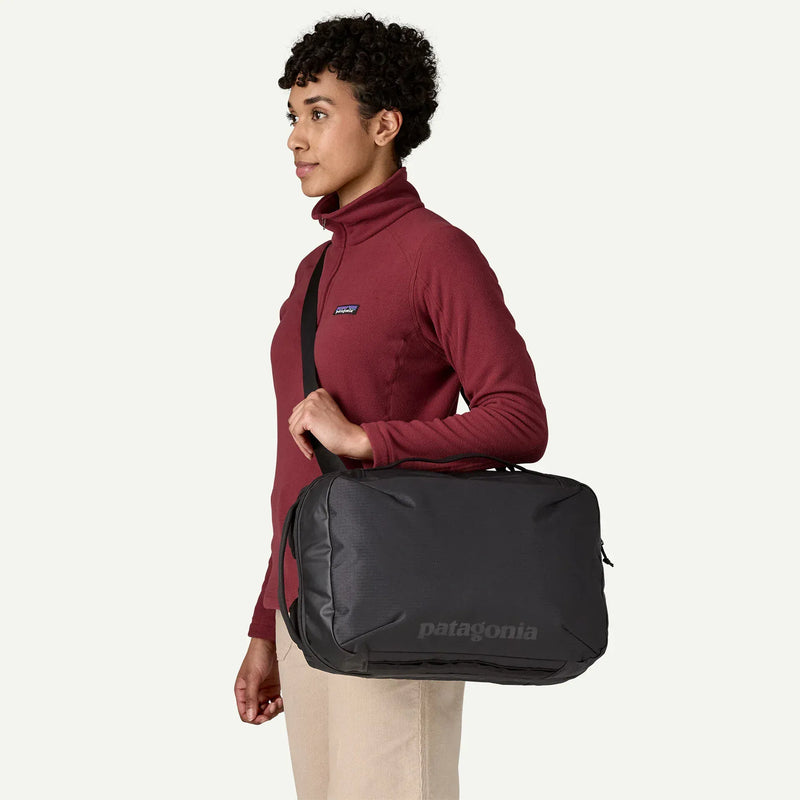 Load image into Gallery viewer, Person wearing a maroon jacket with a Patagonia logo, holding a black patagonia duffel bag on a white background