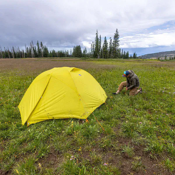 Load image into Gallery viewer, Yellow tent set up in a grassy field with a person sitting nearby under a cloudy sky.