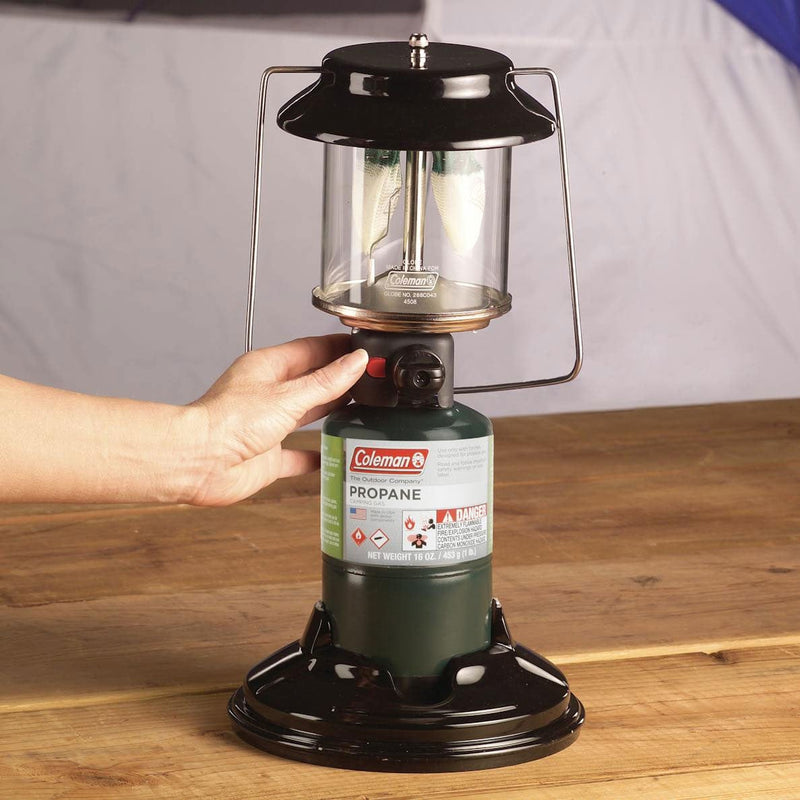 Load image into Gallery viewer, Coleman propane lantern on a wooden surface with a hand holding the fuel canister.
