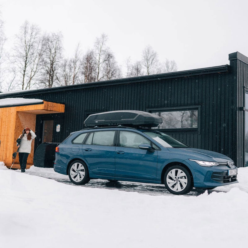 Load image into Gallery viewer, Blue car with a roof box parked in front of a modern building in a snowy landscape.
