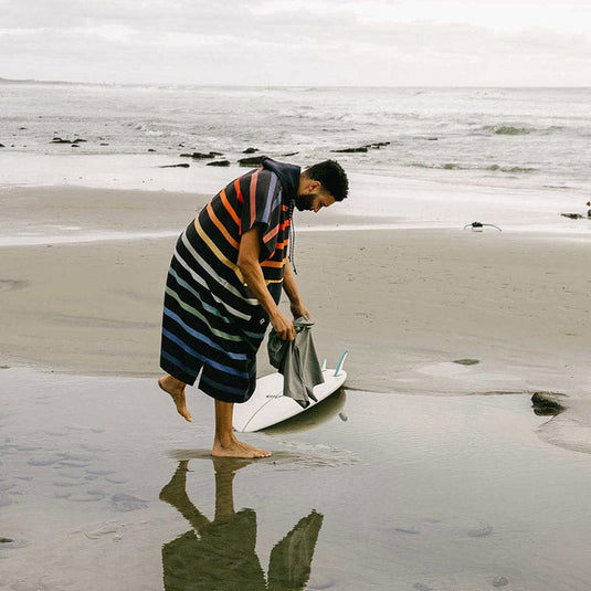 Man on a beach with a striped towel and surfboard, looking at something on the sand.