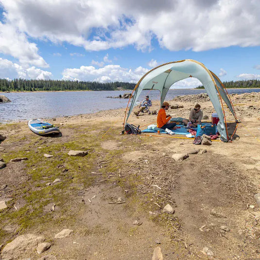 Two people sitting under a tent by a lake with a kayak nearby.