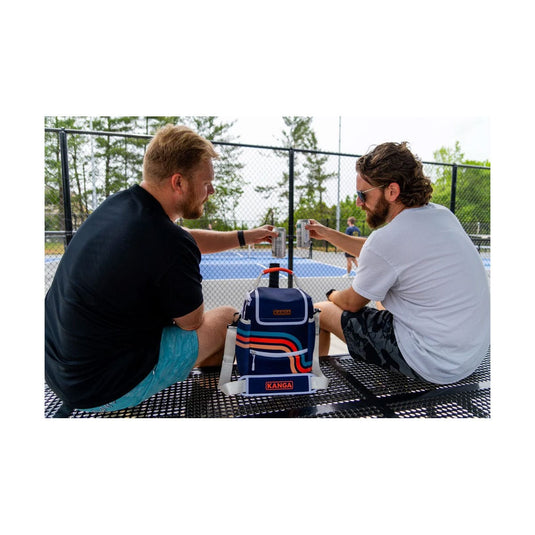 Two men sitting at a table with a cooler labeled 'Kona' on a tennis court.