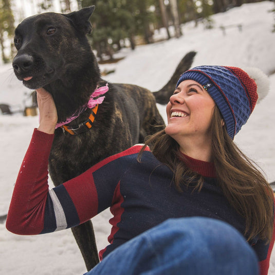 Woman in a blue and red beanie and sweater sitting in the snow with a black dog.