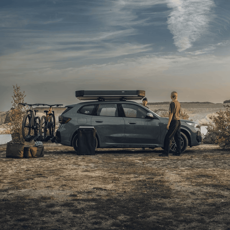 Load image into Gallery viewer, Person standing next to a car with bicycles attached, near a body of water.
