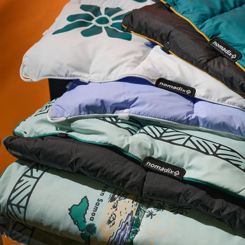 Load image into Gallery viewer, Stack of colorful sleeping bags with 'nomadix' branding on a wooden surface
