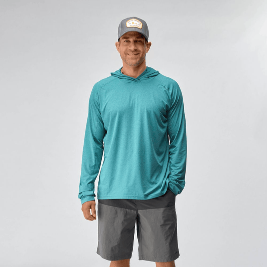 Man wearing a teal long-sleeve shirt, gray shorts, and a cap on a light gray background