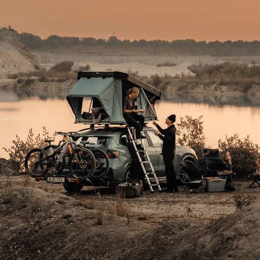 Two people setting up a rooftop tent on a car by a lake at sunset.