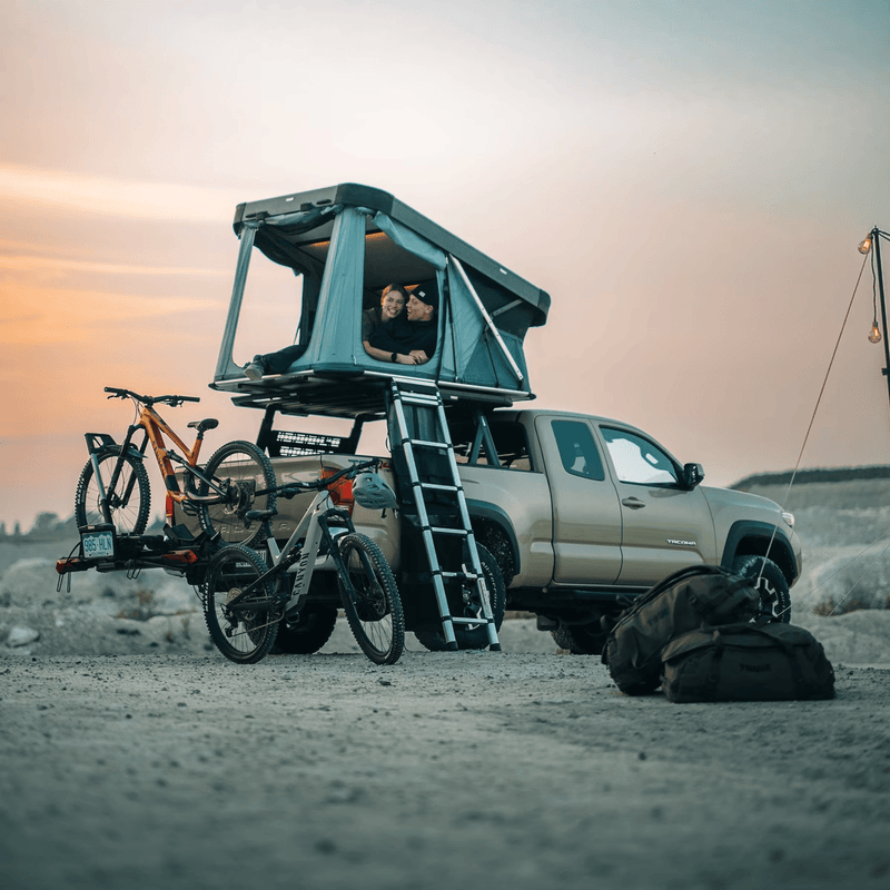 Load image into Gallery viewer, Teal rooftop tent attached to a truck with bicycles on a sandy landscape at sunset.
